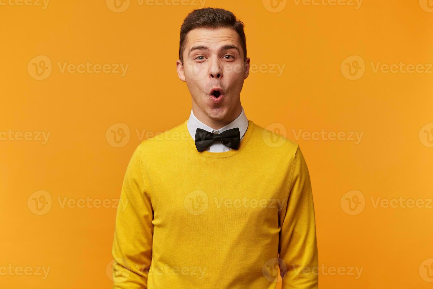 Indoor shot of amazed excited man doesnt believe his success, keeps mouth open, stares at camera, says omg or wow. Pleasant surprised male isolater over yellow background photo