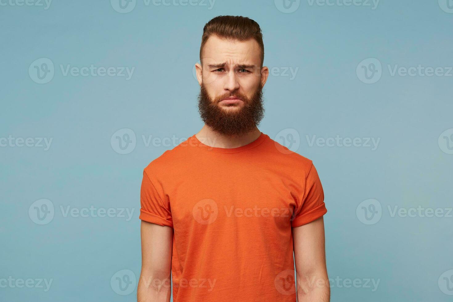 Portrait of emotional bearded man with a sad expression on face, crooked lower lip with a disgruntled look. The male received bad news, feels disappointed, isolated over blue background photo