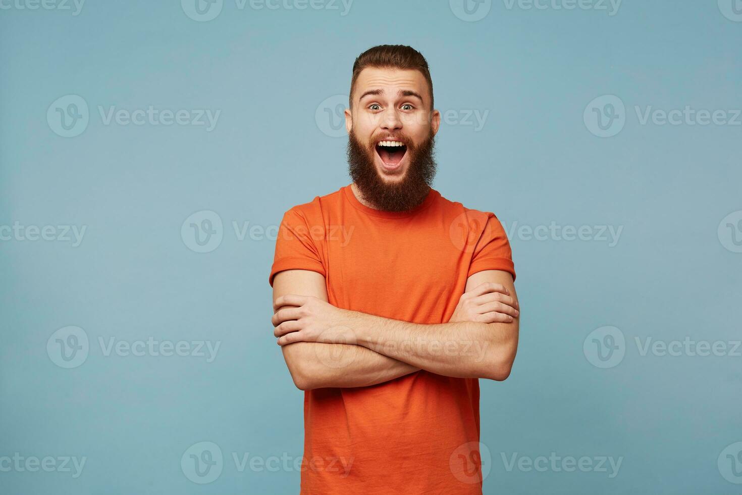 Emotional happy excited funny man with a heavy beard stands with arms crossed and opened mouth in surprisement dressed in red t-shirt isolated over blue background shows wow expression photo