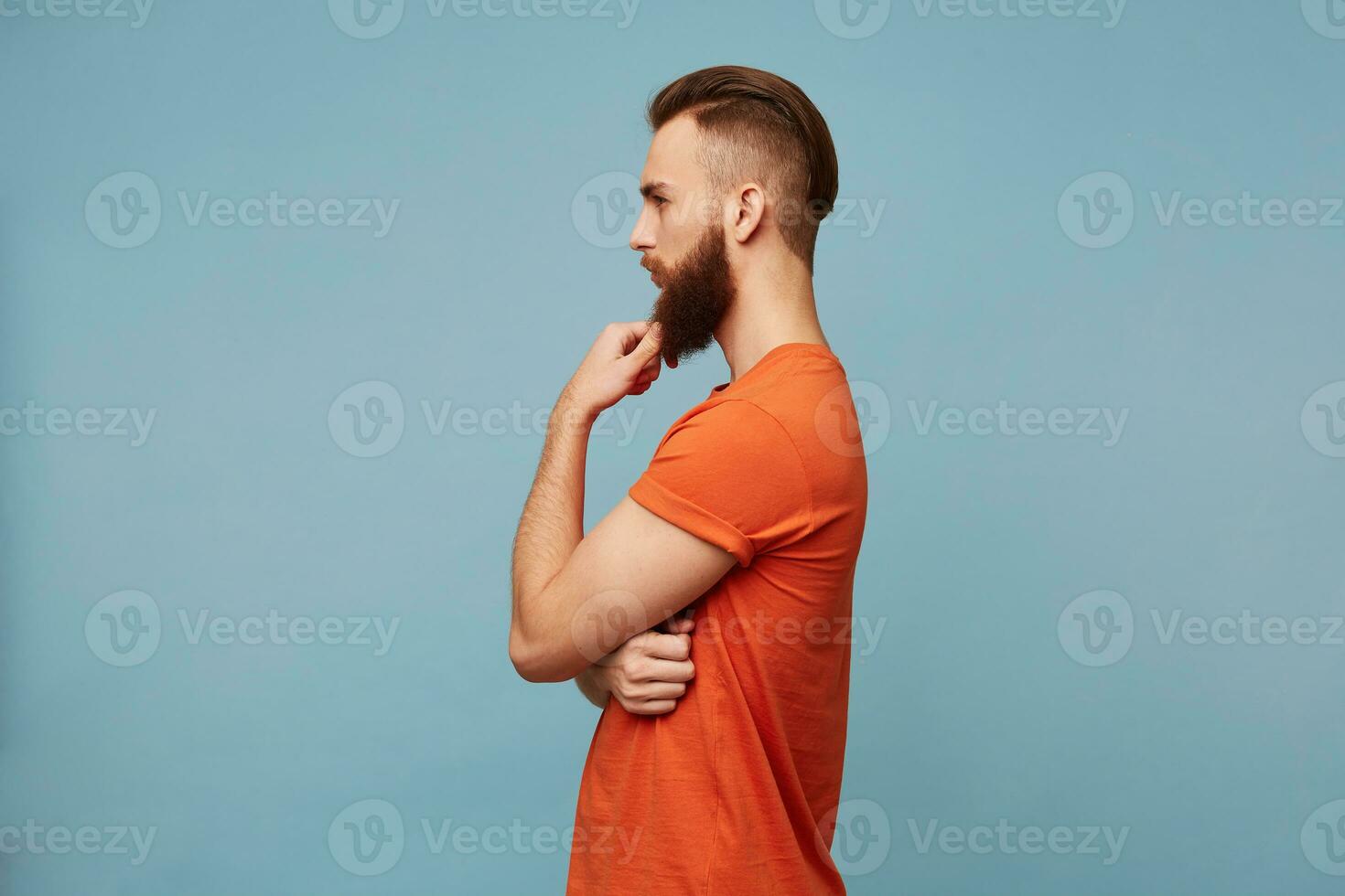 Photo of a young attractive strong man with a fashionable haircut thick beard standing sideways camera dressed in a red T-shirt holds his hand near his chin reflects on something thinks philosophizes.
