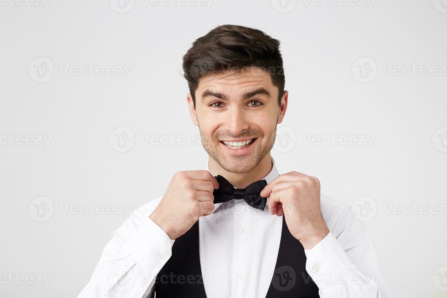 un atractivo encantador moreno es consiguiendo Listo para el noche pelota, vestido en un inteligente traje, endereza un mariposa, sonrisas a su reflexión en el espejo. foto