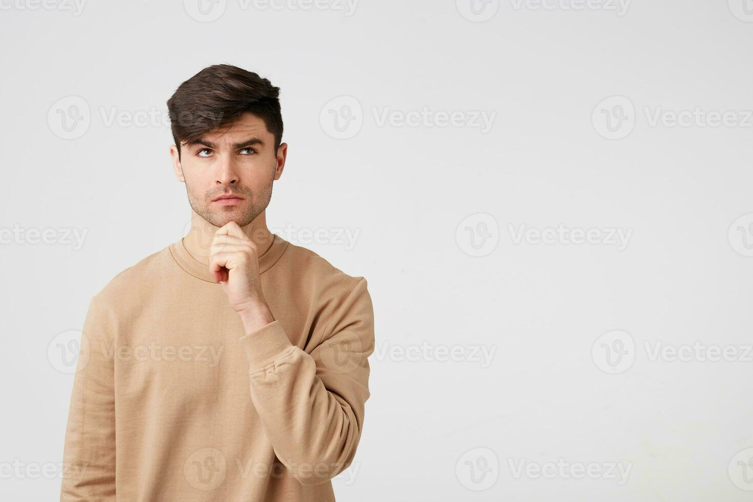 Cute attractive and charming brunet with bristles looks pensive ponders holding a fist near his chin,over white background,looking to the side, standing on the left side,blank copy space for promotion photo