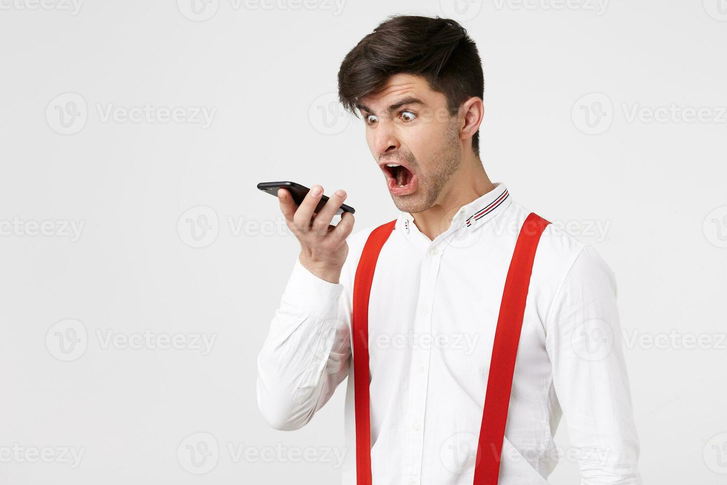 Furious handsome man is screaming on someone via telephone conversation. Guy keeps phone looks at it and yelling with wide opened mouth,wears white shirt and red suspender photo