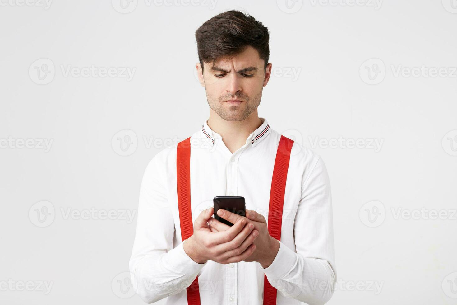 A young guy with a serious thoughtful stressful sad expression looks at the phone that he is holding, chating with someone, feeling dissatisfaction after receiving messages photo