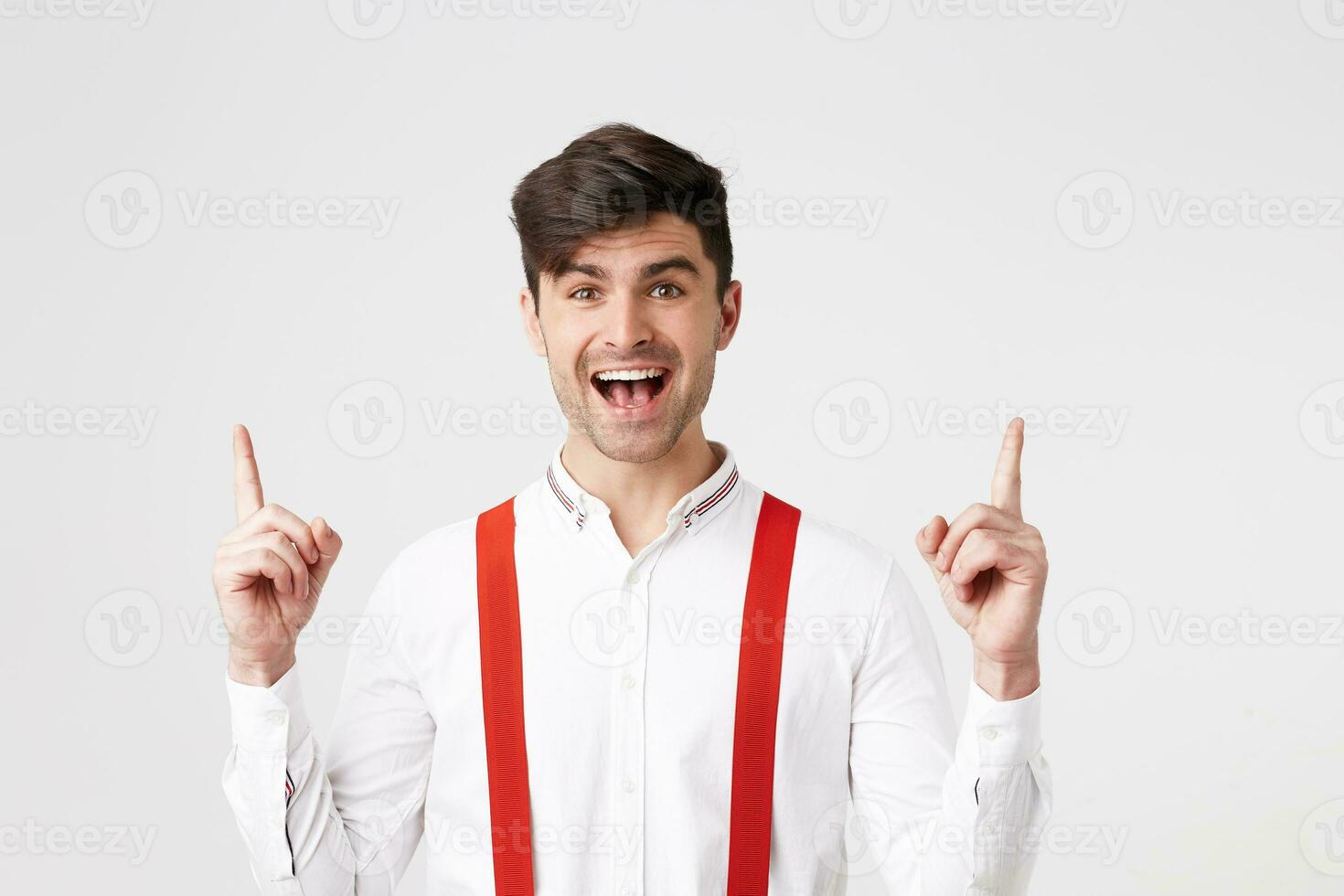 interior foto de contento alegre hermoso chico, mira asombrado emocionado, vestido en blanco camisa y rojo tirantes, puntos con índice dedos hacia arriba en un blanco blanco pared con Copiar espacio tu promocional contenido