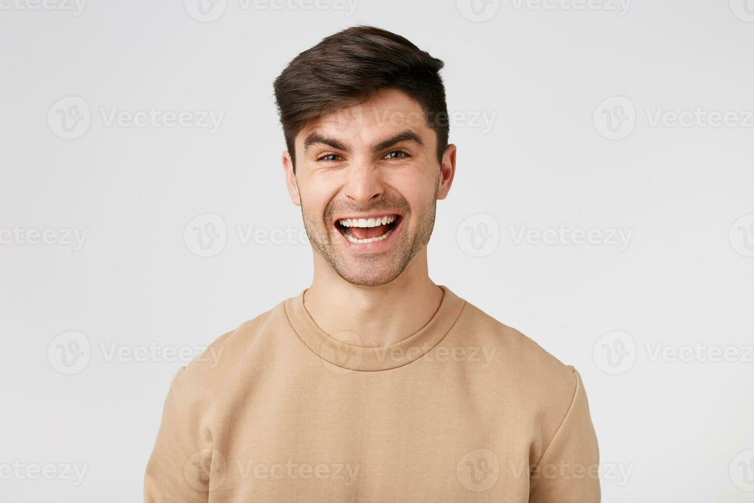 Studio photo of an attractive charming cheerful smiling brunette dressed in nude sweater isolated on a white background. The guy is pleased with his success rejoices in his victory.
