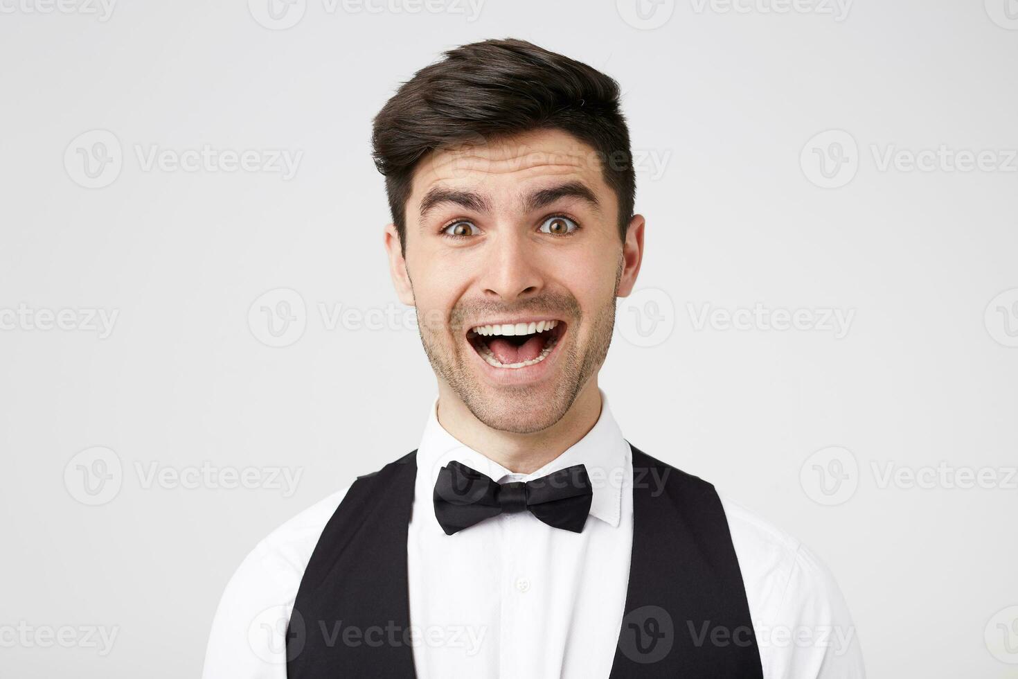 The stylish man fascinated and excited. Close up of the amazed attractive young male in black bowtie looks camera with a toothy surprised smile, standing over white wall photo