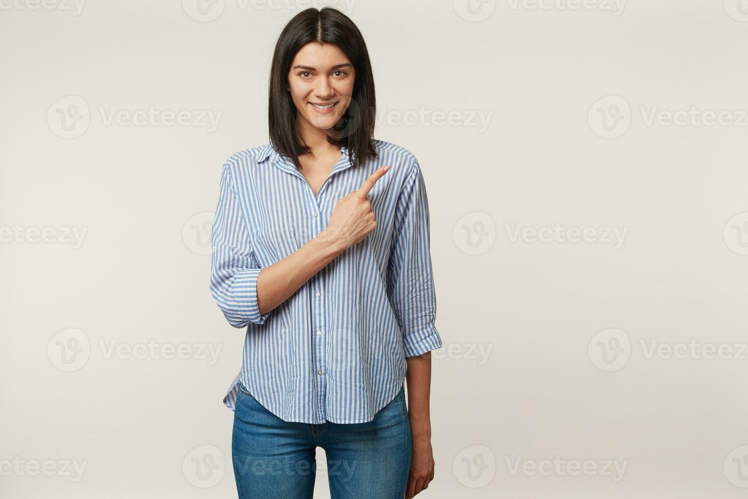 Glad inspired young brunette looks camera with happiness and smile, and pointing index finger there on copy space, dressed in shirt, isolated on a white background photo