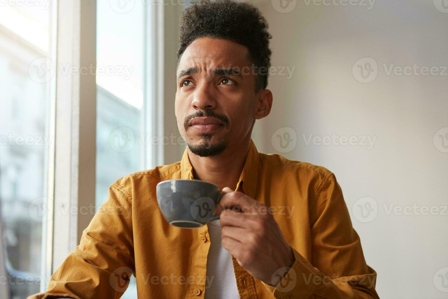 Young thinking African American man wears in yellow shirt, sitting at a table in a cafe and drinks aromatic coffee, a little distressed. Looking thoughtfully into the distance. photo