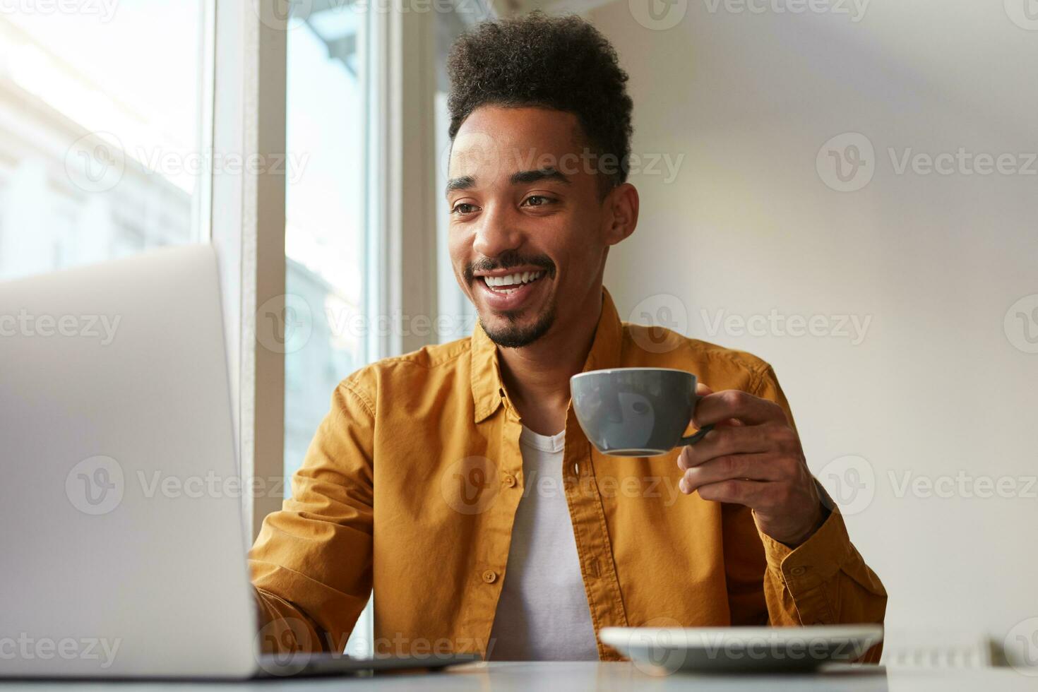 African American guy sitting at a table in a cafe and working on a laptop, wears in yellow shirt, drinks aromatic coffee, chatting with his boss, enjoys the work of a freelancer. photo