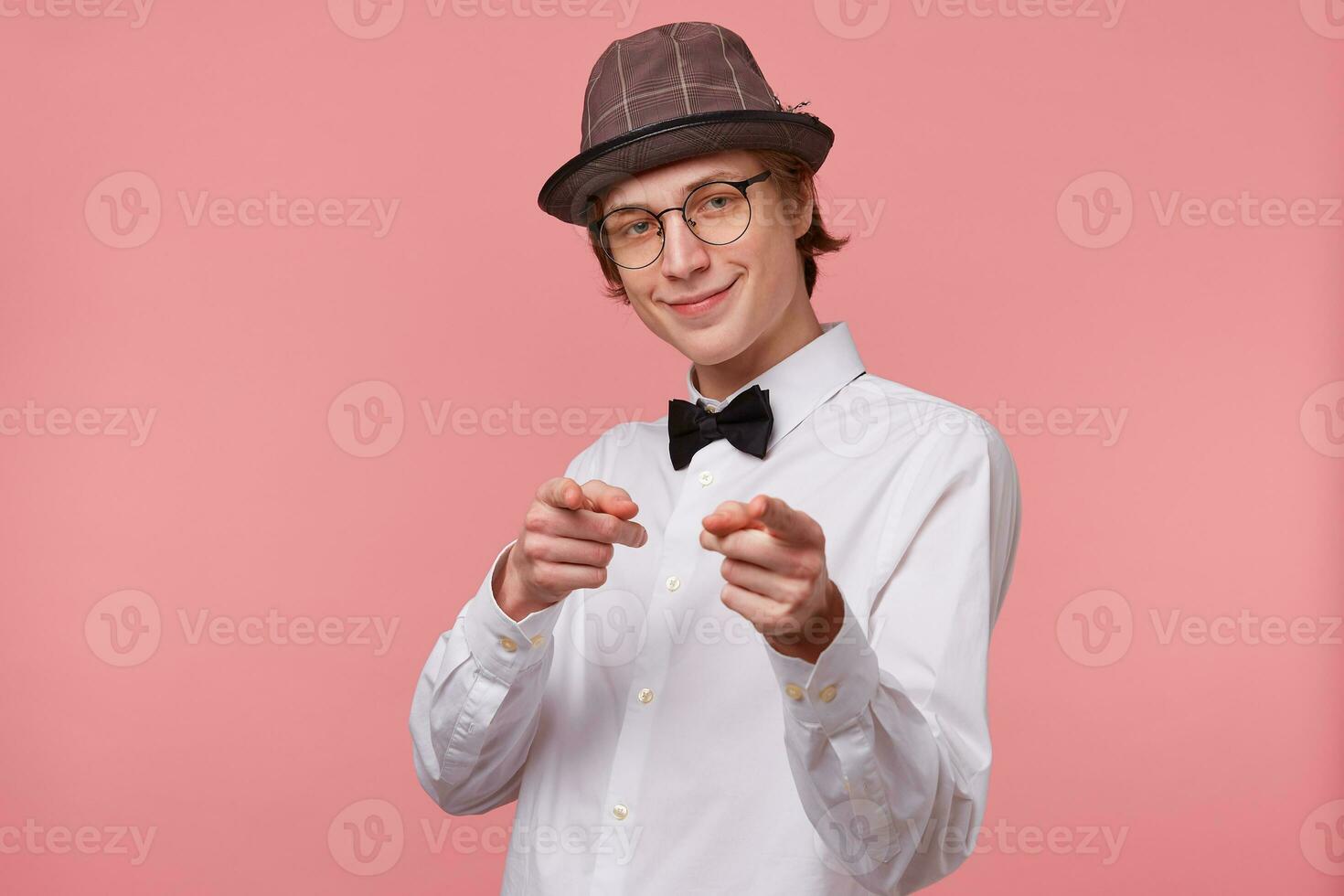 The guy isolated on pink background,dressed in white shirt, hat and black bowtie wears glasses happily pointing with index fingers to a camera like on winner,shows on favorites, with joy and happiness photo