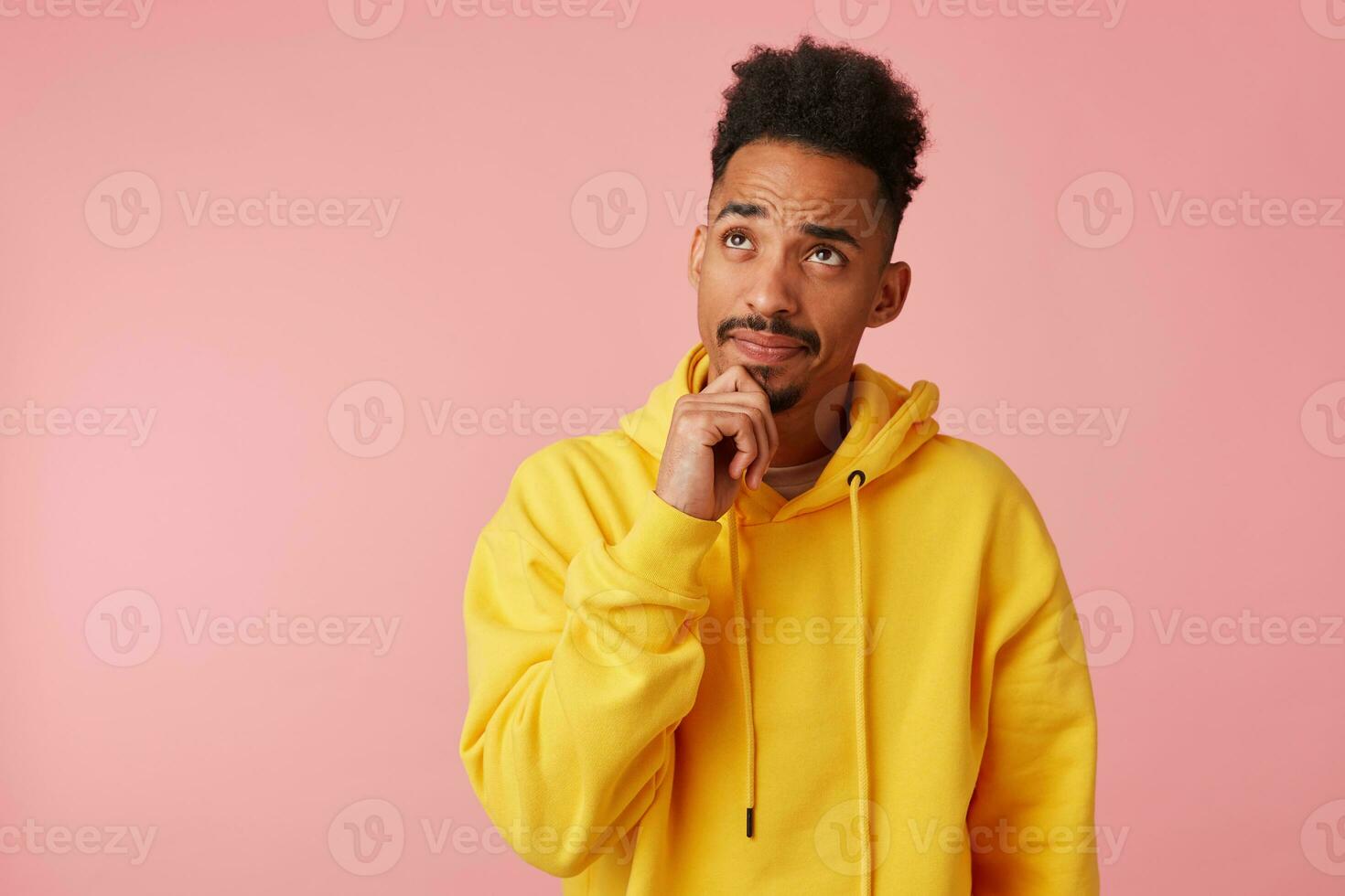 Young thinking african american guy in yellow hoodie, looks up and touches chin, stands over pink background with copy space. photo