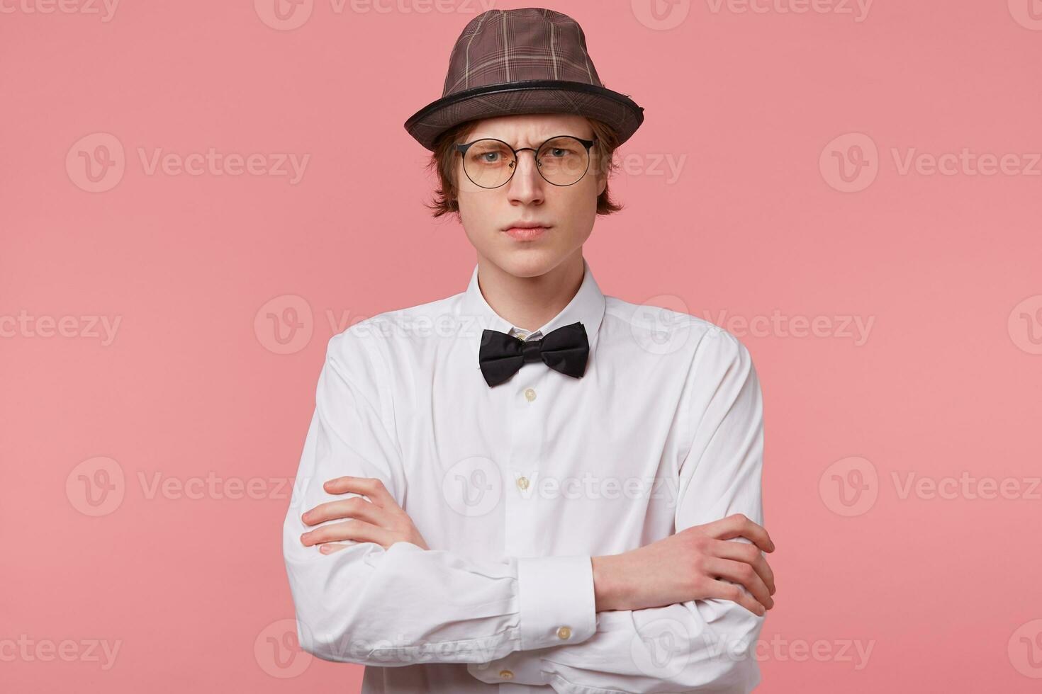 Portrait of serious rude frowning guy in white shirt, hat and black bowtie wears glasses looks angry, standing with arms crossed, isolated on pink background photo