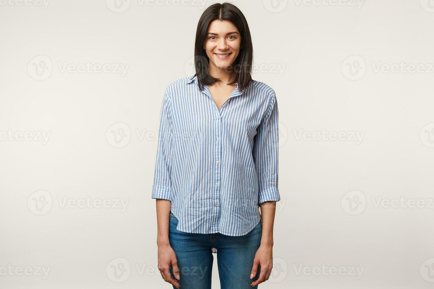 Attractive pleasant young dark-haired female standing dressed in jeans and striped shirt. Charming nice positive girl isolated over white background photo