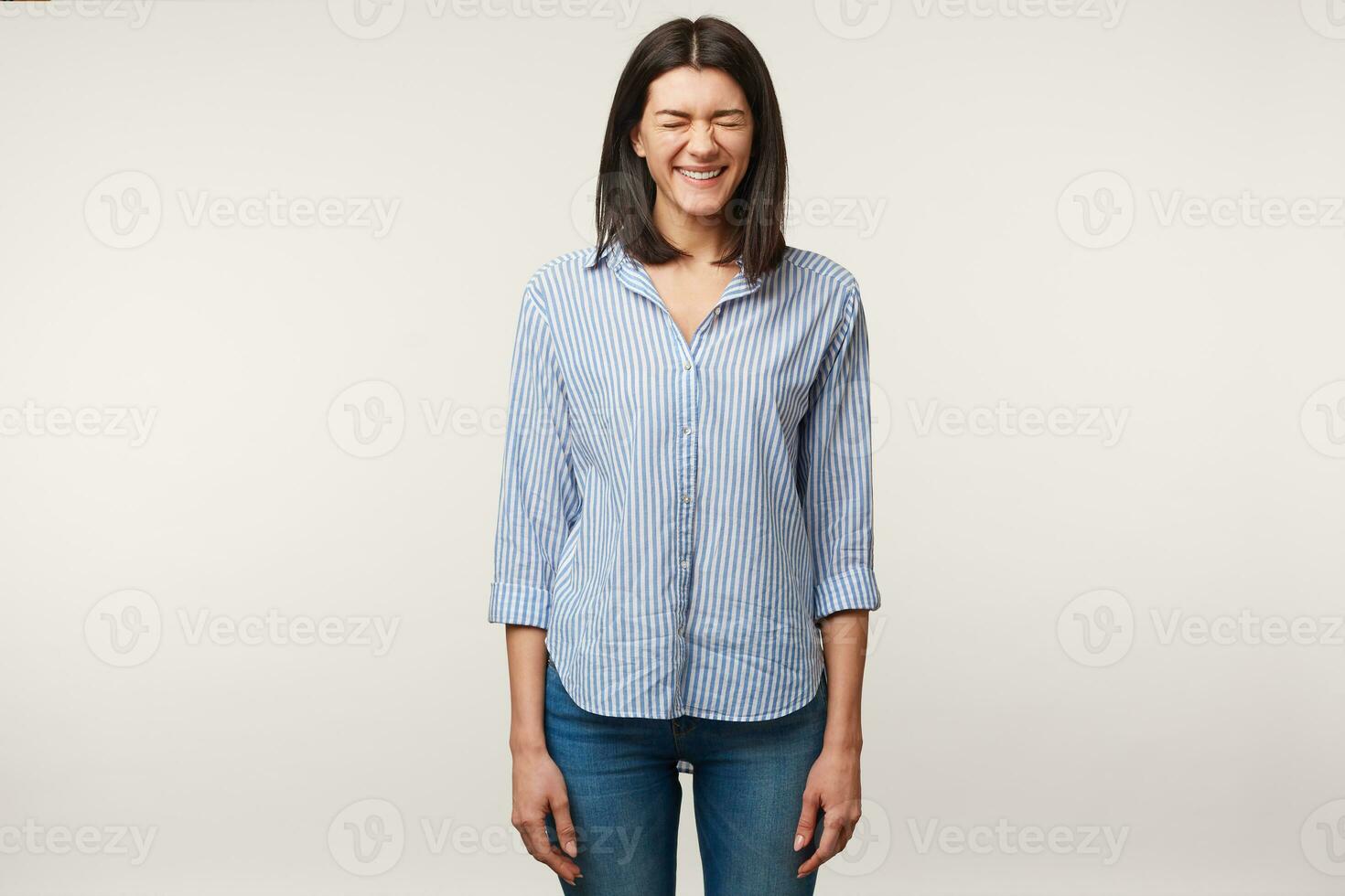 Attractive pleasant young dark-haired female standing dressed in jeans and striped shirt waiting for some unexpected or astonishing surprise from boyfriend with closed eyes isolated over white wall photo