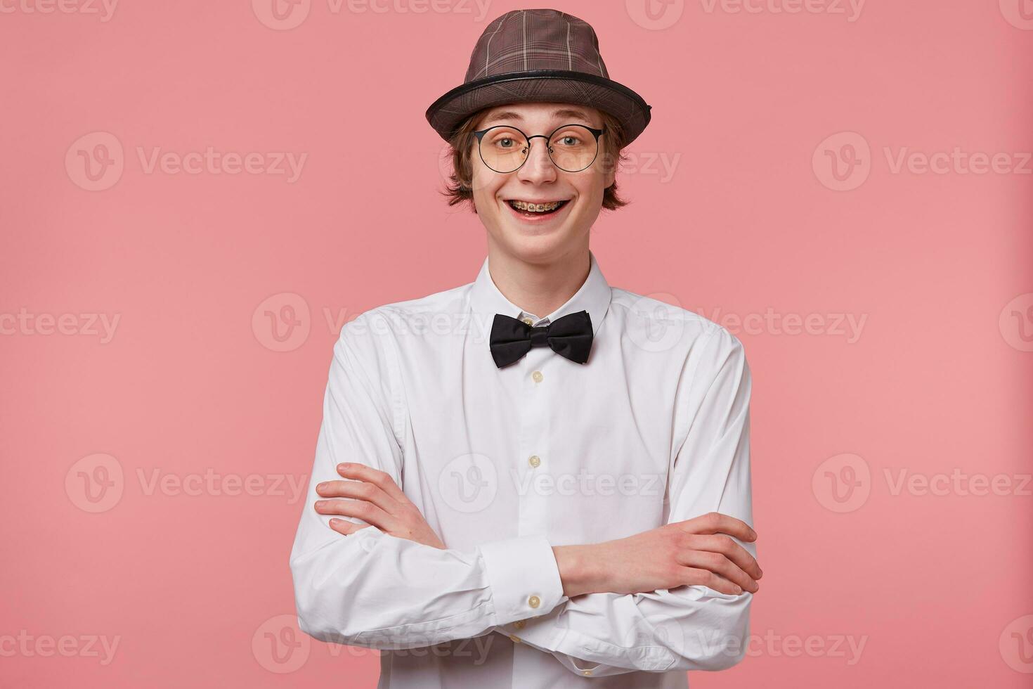 Portrait of joyful funny young guy in white shirt, hat and black bowtie wears glasses happily laughing showing orthodontic brackets,standing with hands crossed, isolated on pink background photo
