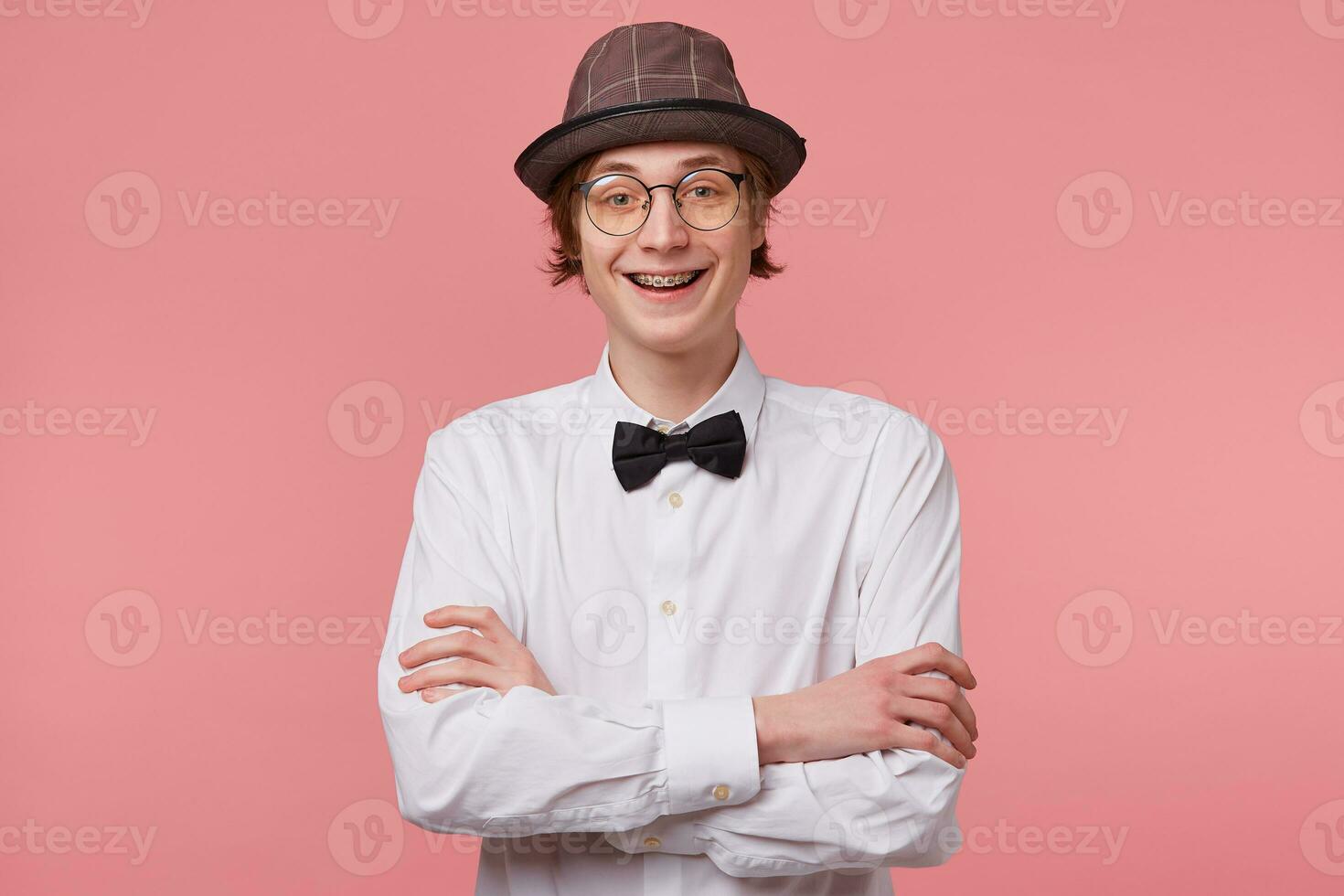 Horizontal portrait of joyful nice young guy in white shirt, hat and black bowtie wears glasses happily smiling showing braces, standing with hands crossed, isolated on pink background photo