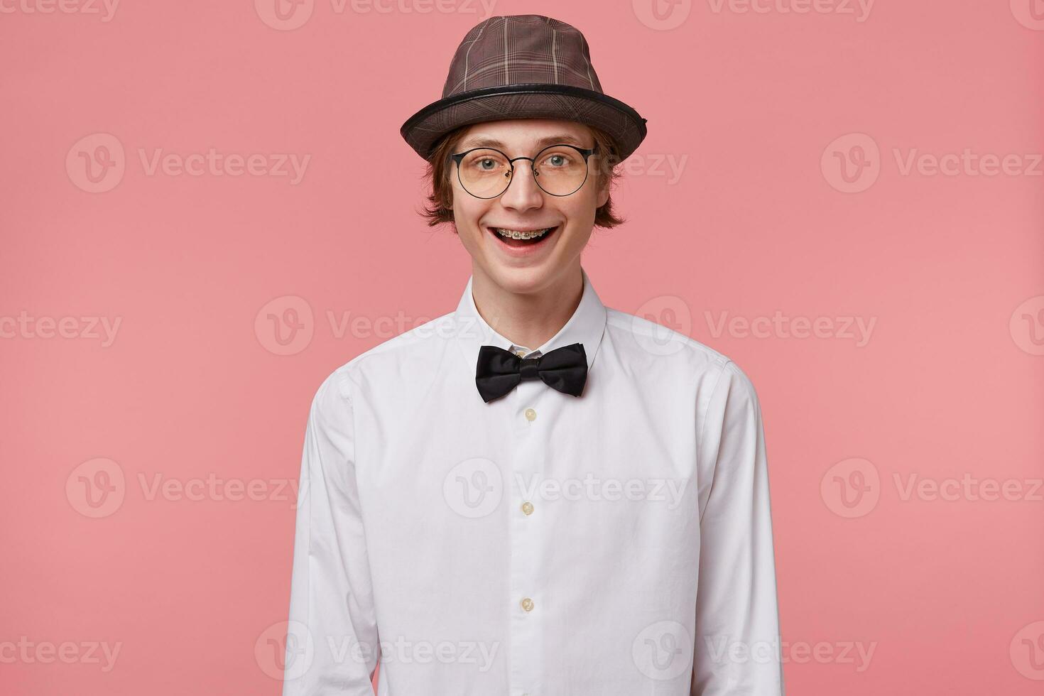 Nice young guy in white shirt, hat and black bowtie wears glasses happily widely smiling showing orthodontic brackets, isolated on pink background photo