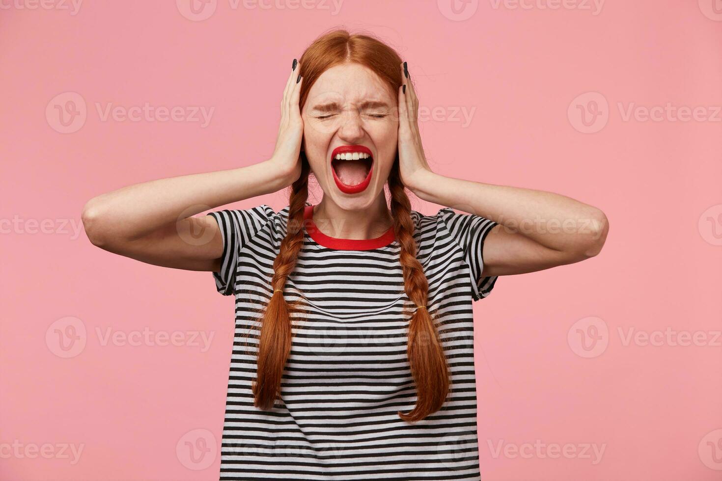 Angry young red-haired girl in stripped t-shirt, keeps closed eyes, closes her ears with palms, showing gesture of ignore, demonstrates loud scream or shout, over pink wall photo