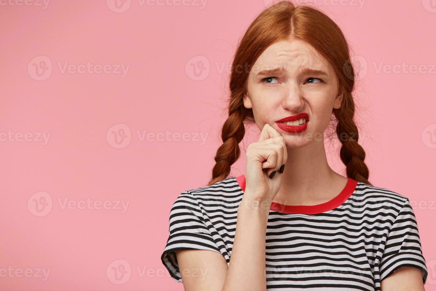 Dissatisfied grumpy unsatisfied frowning red-haired girl with two braids keeps fist near chin and looking to the upper left corner with discontent, isn't thrilled with new idea, on pink background photo