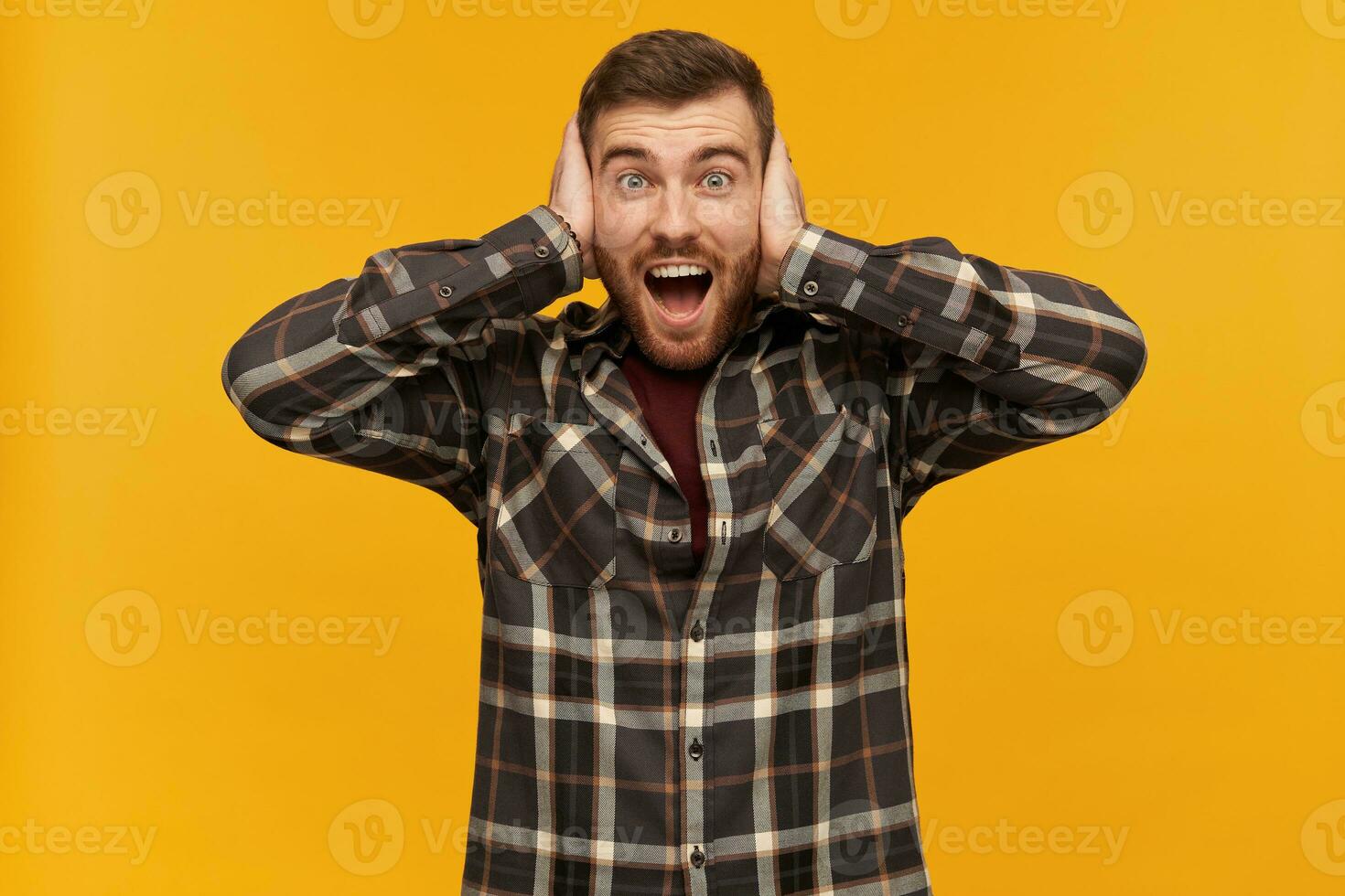 Bearded guy, happy looking man with brunette hair. Wearing checkered shirt and accessories. Emotion concept. Cover ears with palms. Watching excited at the camera, isolated over yellow background photo