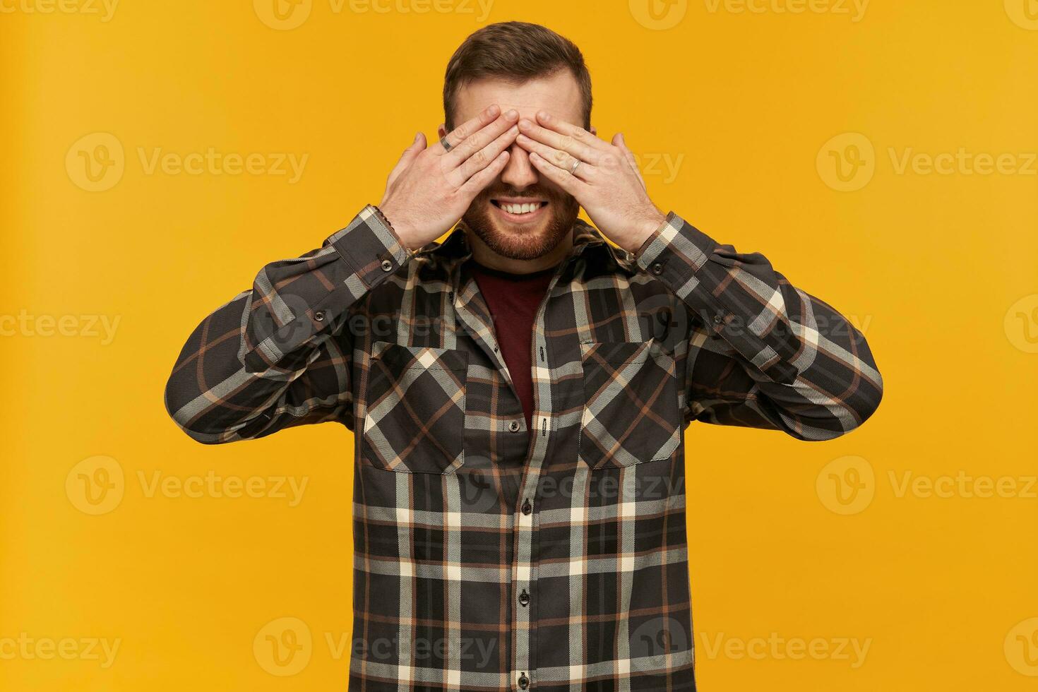 Bearded smiling guy, happy looking man with brunette hair. Wearing checkered shirt and accessories. Close his eyes with palms. Hide and seek. Stand isolated over yellow background photo