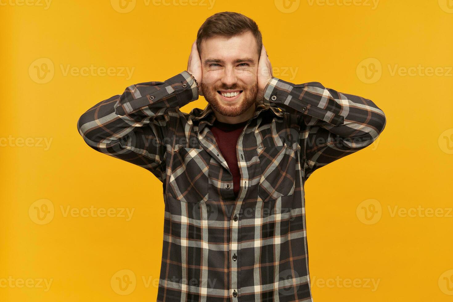 Portrait of handsome, joyful male with brunette hair and bristle. Wearing checkered shirt and accessories. Cover ears with palms. Can't hear. Watching at the camera, isolated over yellow background photo