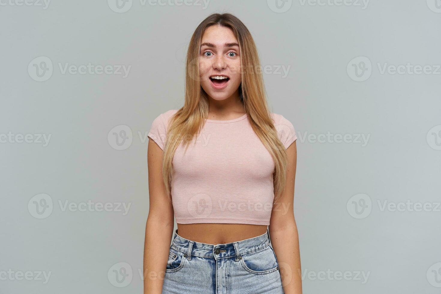 Portrait of confused, disturbed girl with blond long hair. Wearing pink t-shirt and jeans. People and emotion concept. Watching at the camera isolated over grey background photo