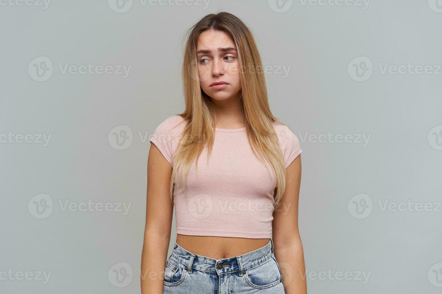 Sad looking woman, disappointed girl with long blond hair. Wearing pink t-shirt and jeans. People and emotion concept. Watching to the left at copy space, isolated over grey background photo