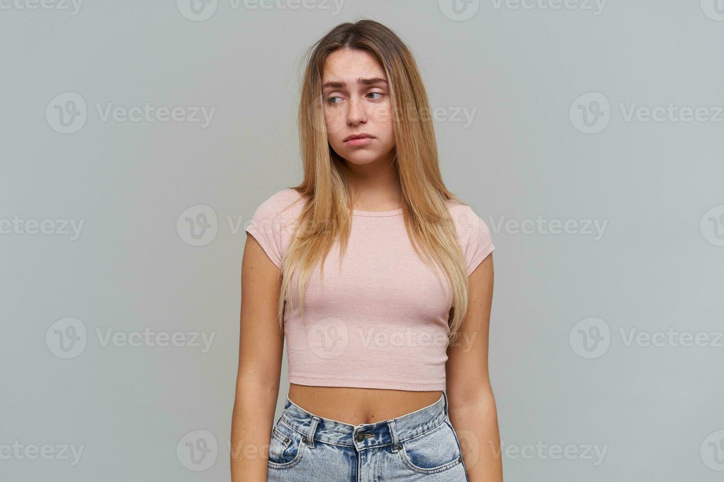 Sad looking woman, disappointed girl with long blond hair. Wearing pink t-shirt and jeans. People and emotion concept. Watching to the left at copy space, isolated over grey background photo