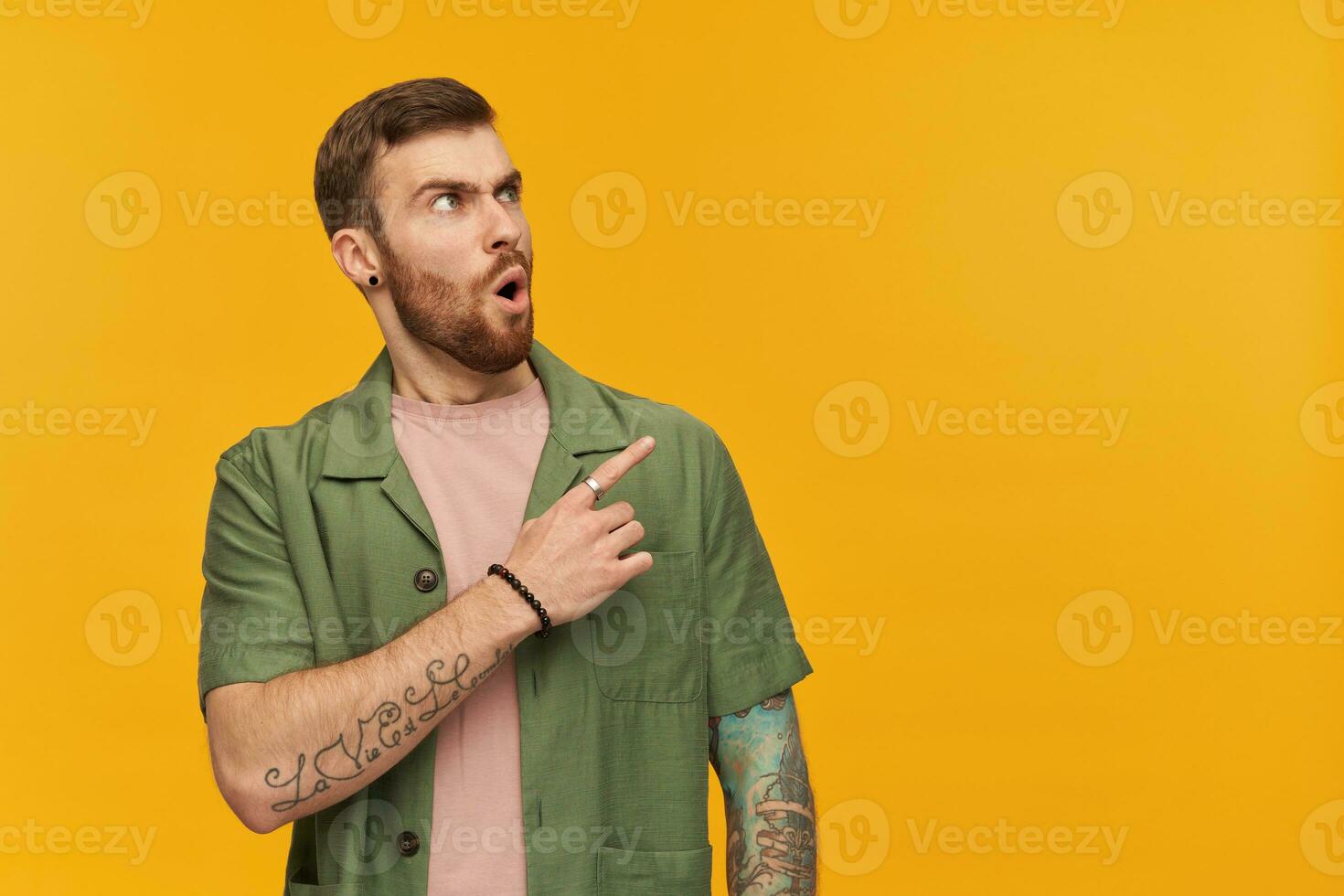Shocked male, frowning guy with brunette hair and beard. Wearing green short sleeves jacket. Has tattoo. Watching and pointing finger to the right at copy space, isolated over yellow background photo