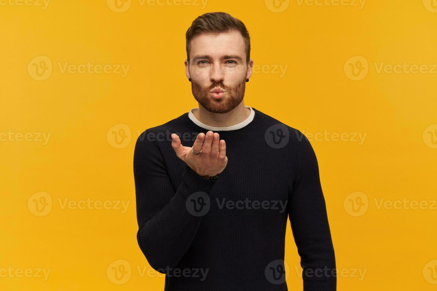 Bearded attractive man, flirty guy with brunette hair. Has piercing. Wearing black sweater. Purses lips and sending air kiss. Held out palm. Watching at the camera, isolated over yellow background photo