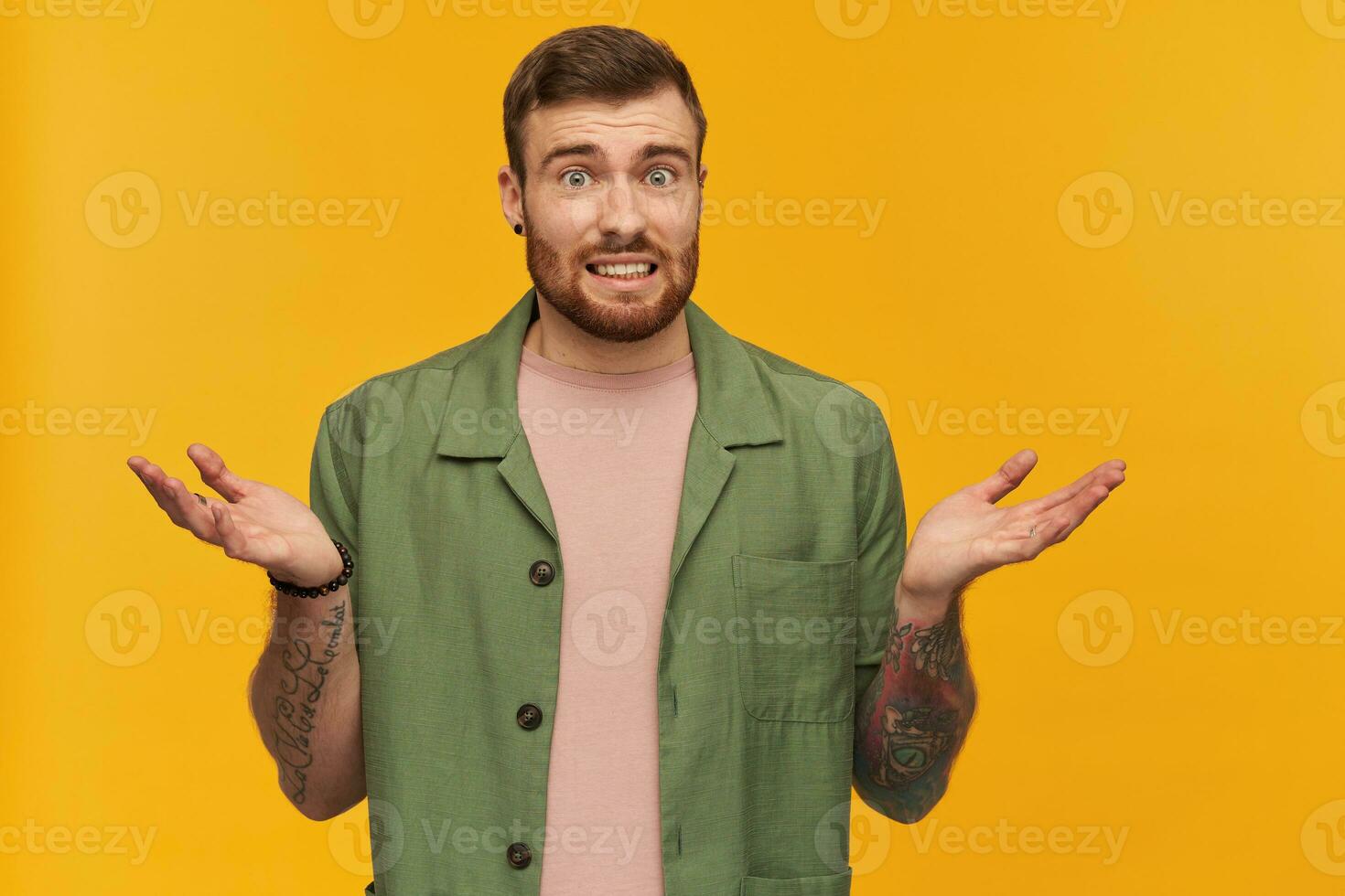 Bearded guy, bewildered looking man with brunette hair. Wearing green short sleeves jacket. Has tattoo. Shrugs and shows unsure gesture. Watching at the camera, isolated over yellow background photo