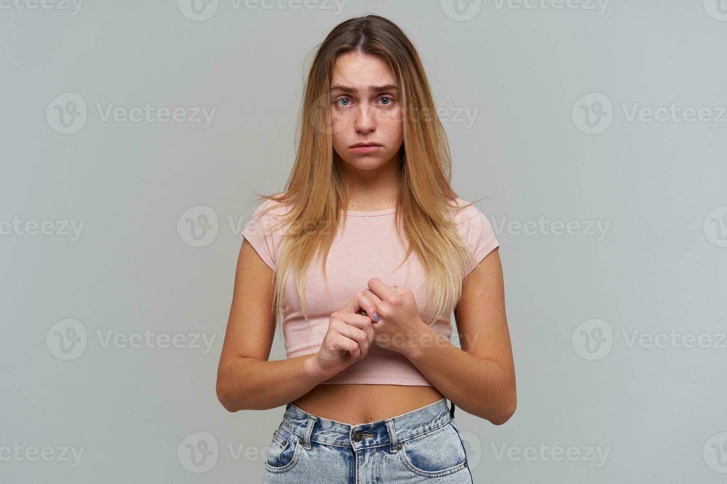 Unhappy girl, disappointed woman with blond long hair. Wearing pink t-shirt and jeans. People and emotion concept. Nervously touching fingers. Watching at the camera isolated over grey background photo