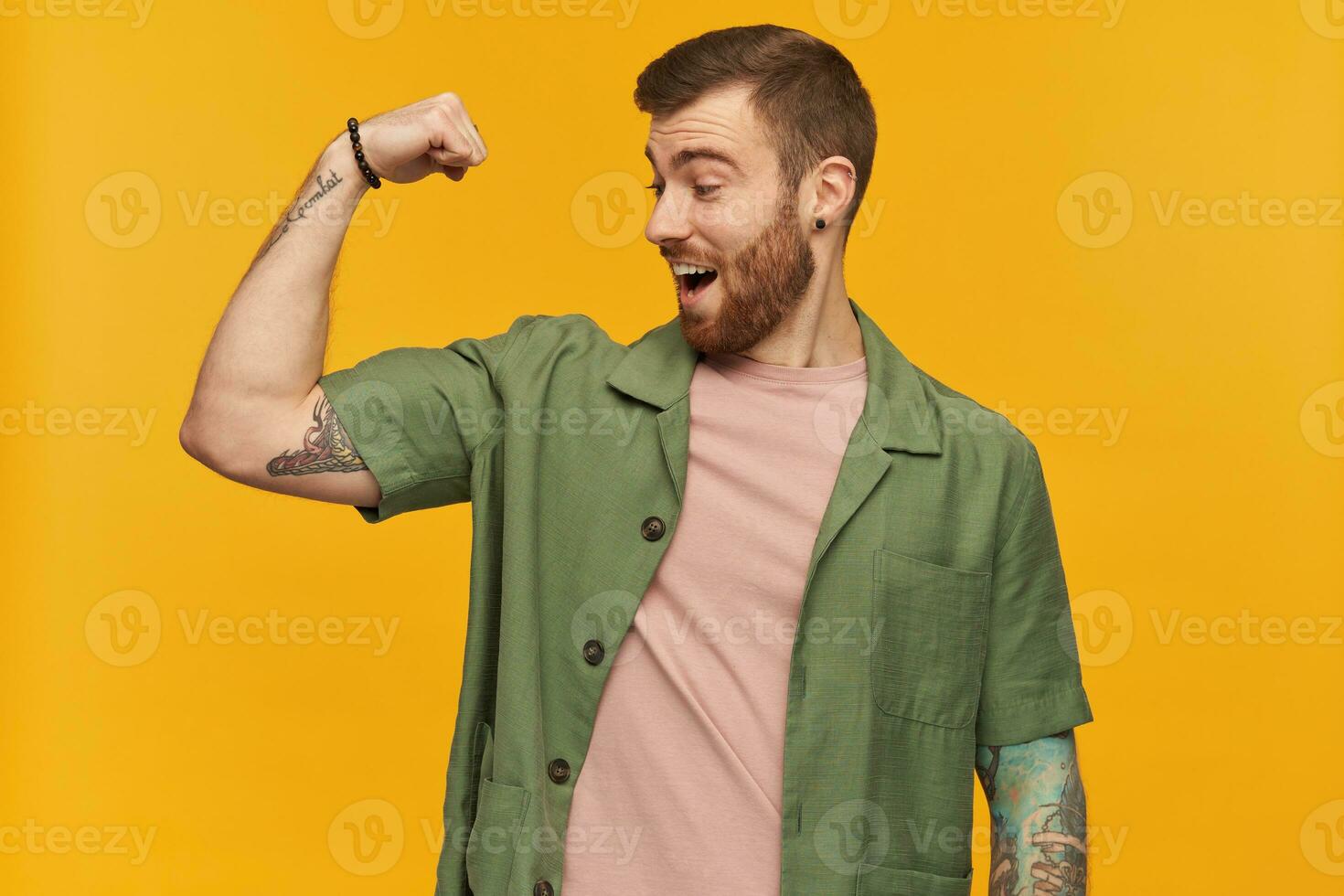 Portrait of handsome male with brunette hair and bristle. Wearing green short sleeves jacket. Has tattoos. Showing his biceps and watching at it excited. Stand isolated over yellow background photo