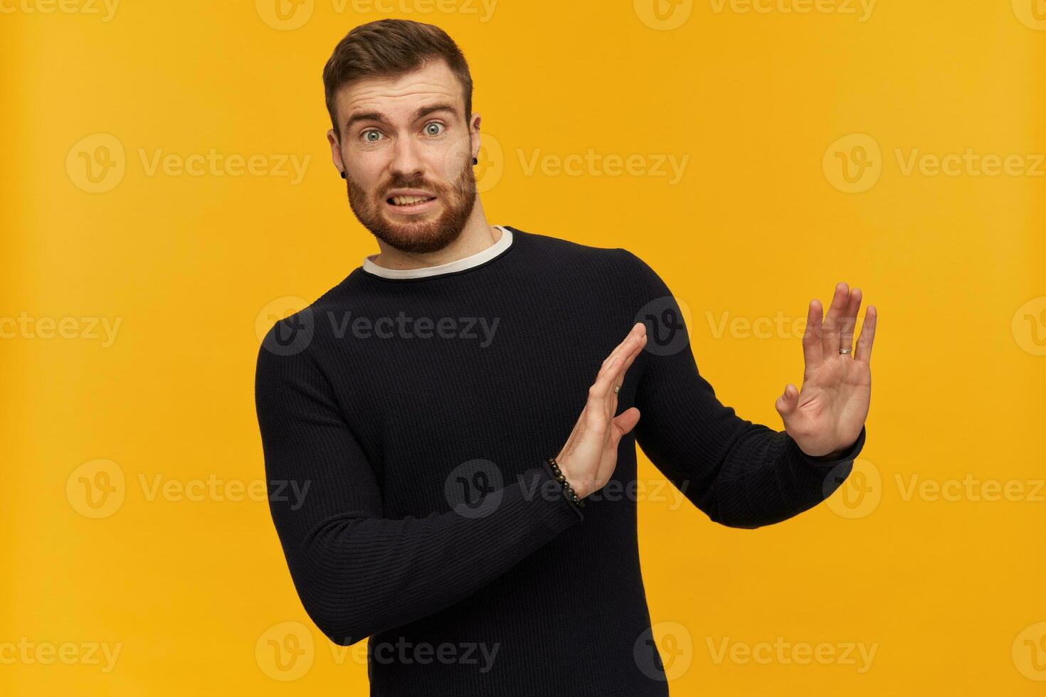 Bearded shocked guy, skeptical looking man with brunette hair. Has piercing. Wearing black sweater. Pushes arms away, reject gesture. Watching at the camera, isolated over yellow background photo
