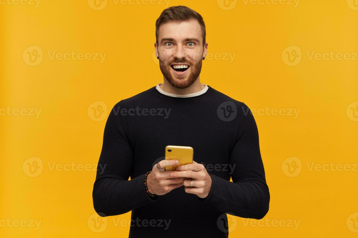 Portrait of cheerful, smiling male with brunette hair and beard. Has piercing. Wearing black sweater. Holding a smartphone. Watching excited at the camera, isolated over yellow background photo