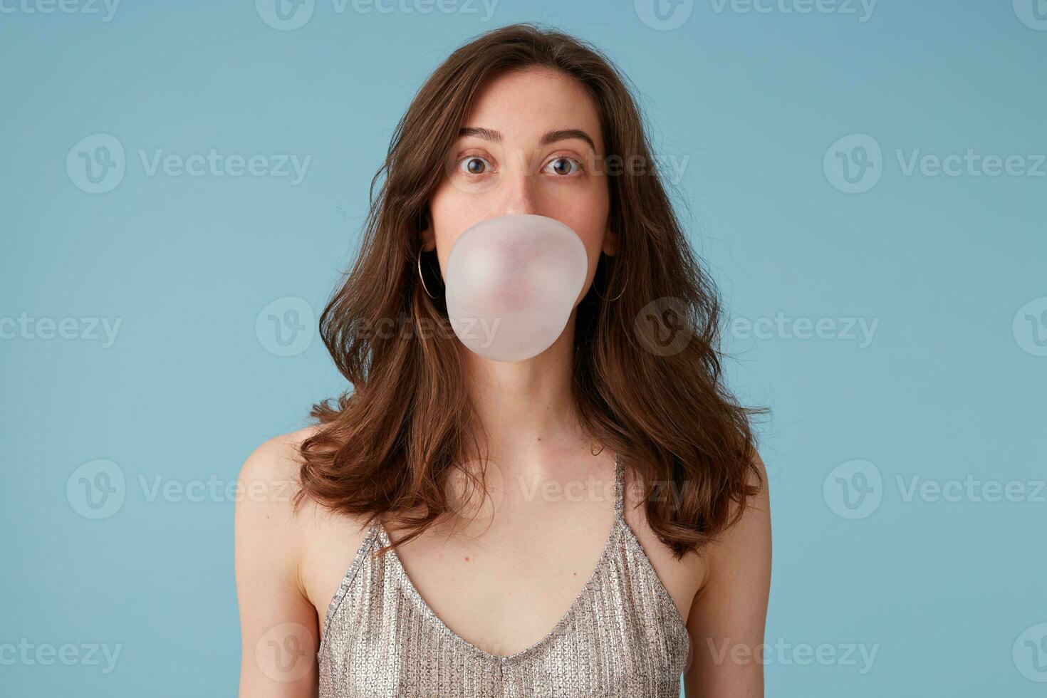 retrato de bonito niña inflar un burbuja, masticación un chicle, teniendo divertido, en un fiesta vestido, aislado terminado azul antecedentes foto