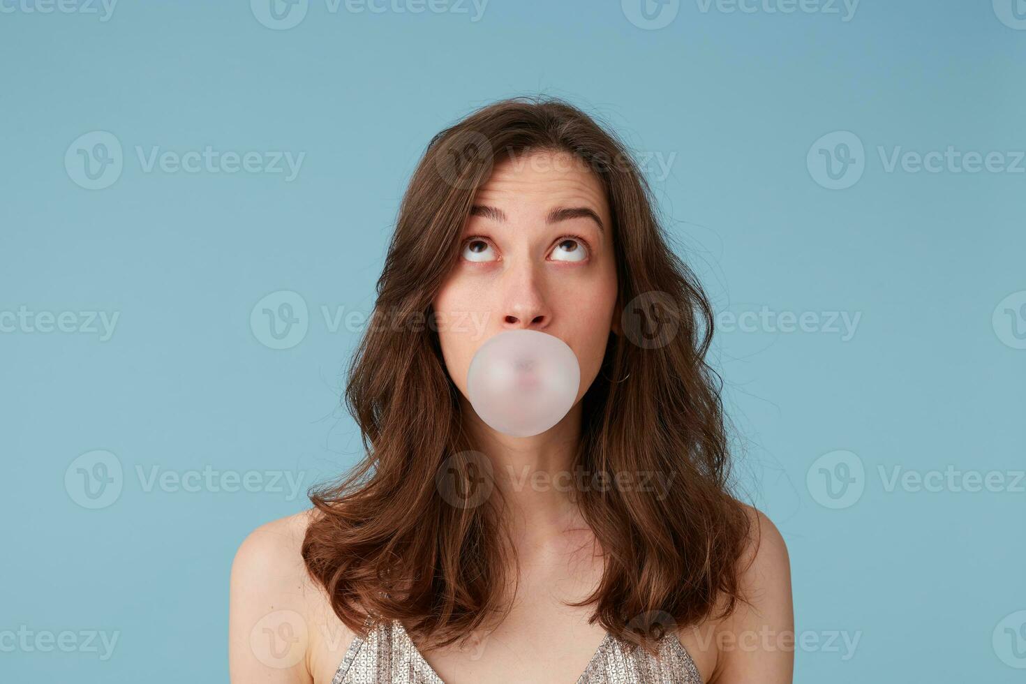 Looking upwards on a blank copy space amazed nice girl inflating a bubble, chewing a gum, in a party dress, isolated over blue background photo