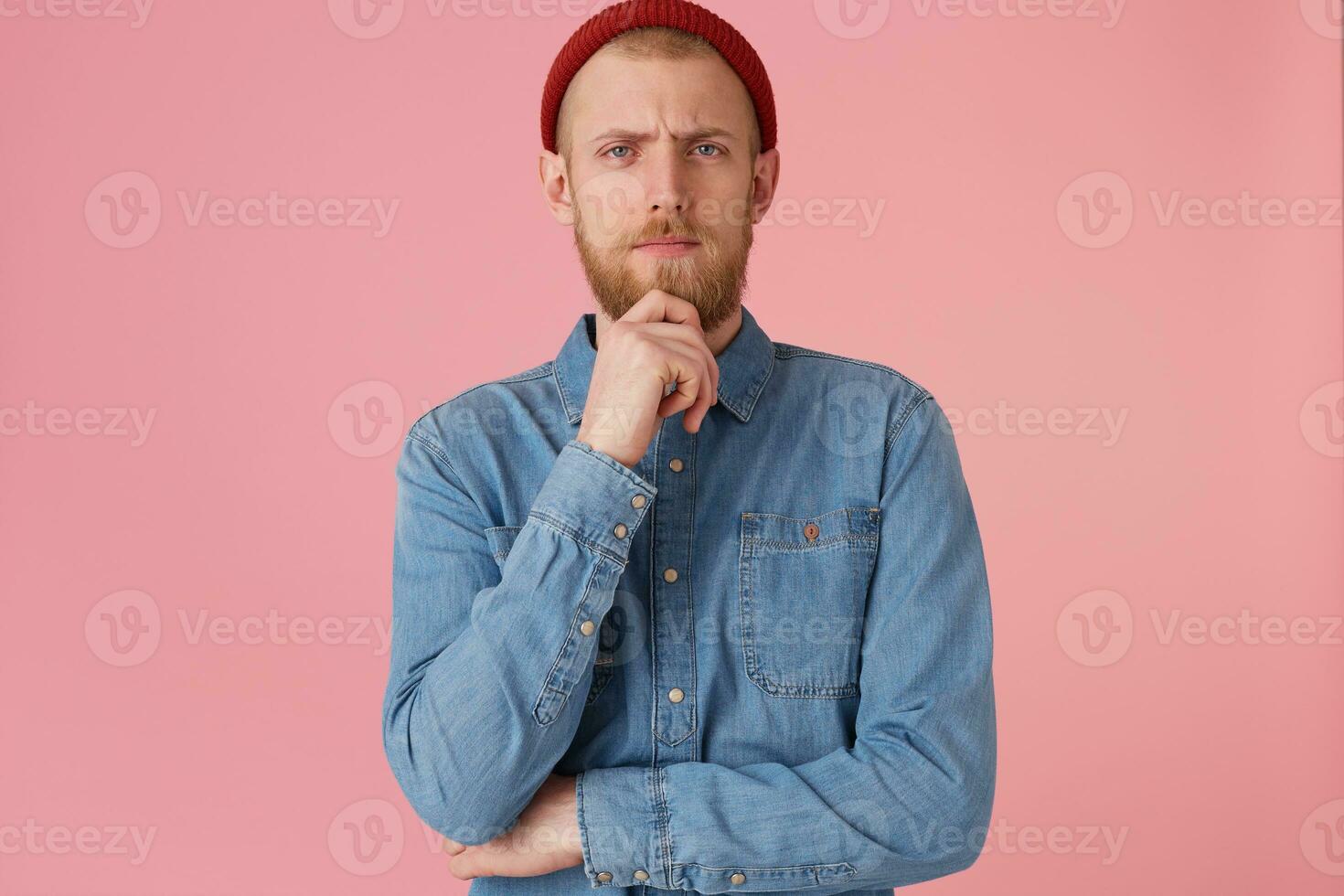 A handsome thoughtful bearded guy looking camera holding his chin, thinks about his future, make plans, dreams, feels confident in his abilities, isolated over pink background photo