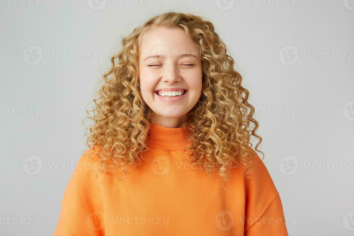 Smiling blonde with curly hair, stands contentedly closed her eyes and rejoices, enjoys the moment, imagines something beautiful, dreams, isolated on a white background to wear a warm orange sweater photo