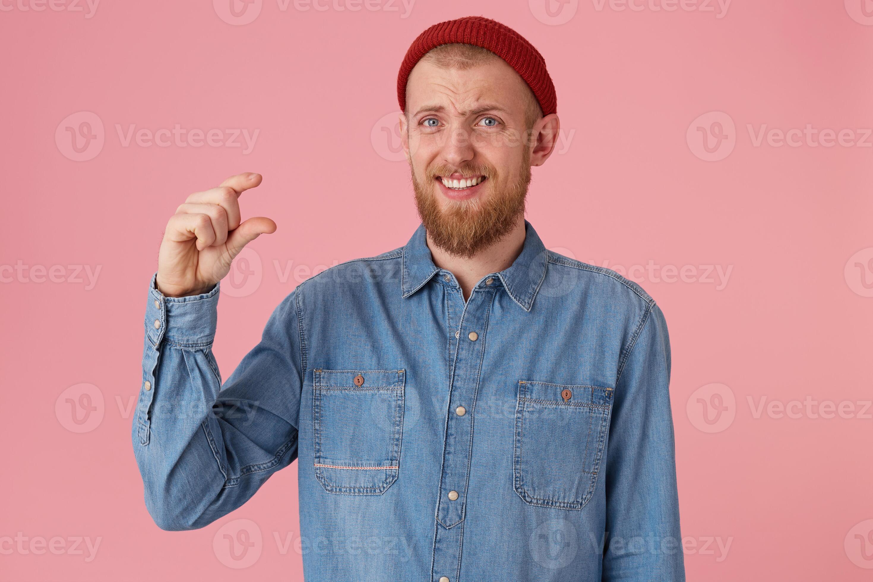 Sckeptical bearded male in funny red hat wears denim shirt, makes a