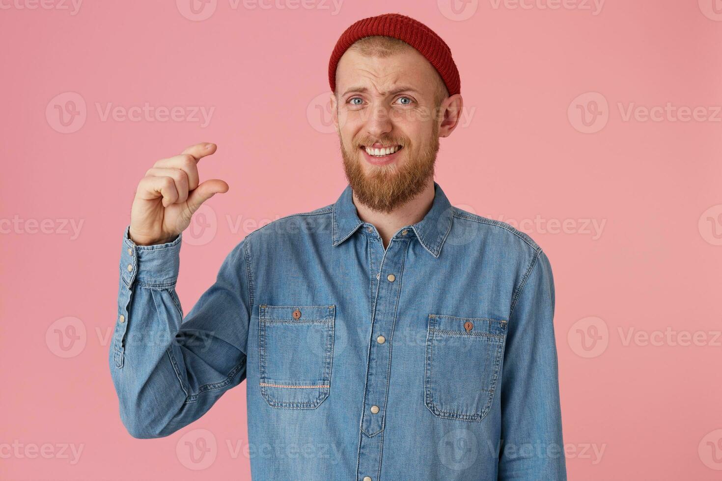 Sckeptical bearded male in funny red hat wears denim shirt, makes a hand gesture, demonstrates something very tiny, poses against pink wall. Puzzled male student shows how much he learned photo