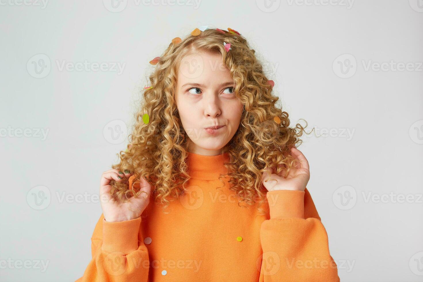 Horizontal shot of attractive young female with pensive contemplating expression,ponders about something,keeps touches her blond hair,dressed in casual orange sweater, isolated over white background photo