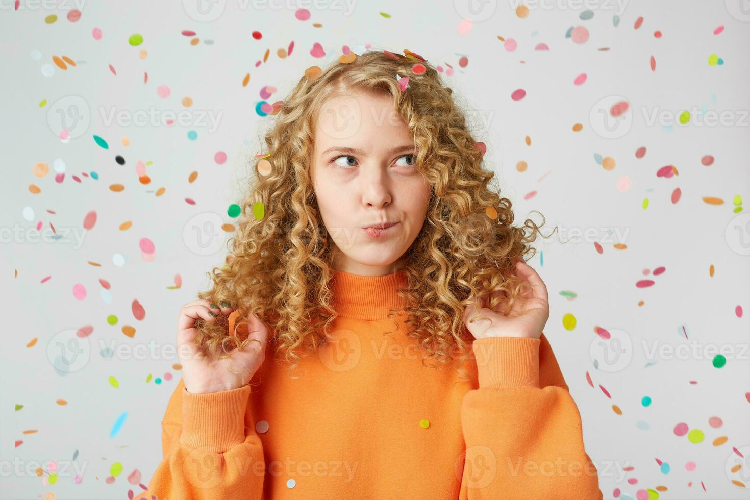 Horizontal shot of attractive young female with pensive contemplating expression,ponders about something,keeps touches her blond hair,dressed in casual orange sweater, stands under falling confetti photo
