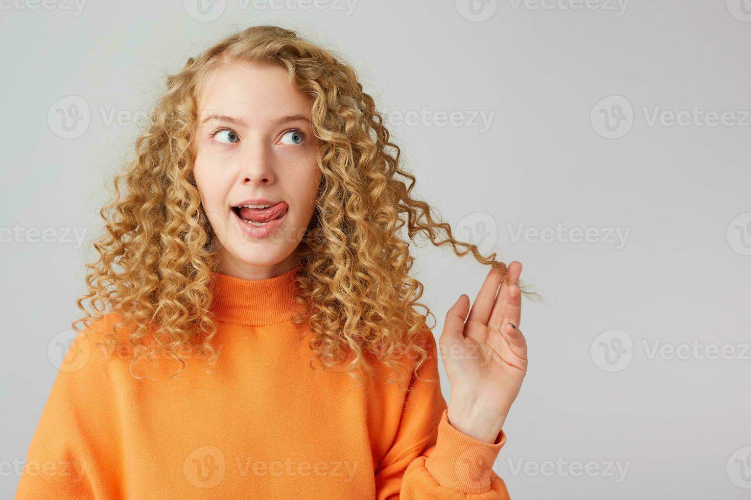 Attractive cute curly blonde in orange sweater dreamy pensively looking towards empty copy space, shows her tongue holding in her hand a strand of hair isolated on a white background photo