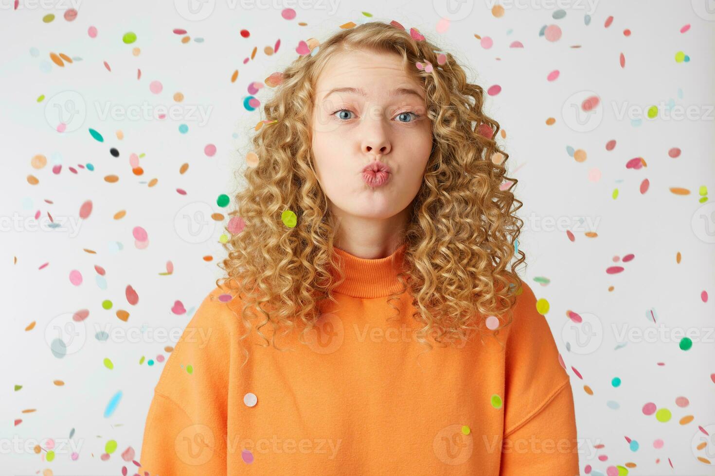 Close up of lovely sweet nice blonde girl sending air kiss directly to the camera with tender look, stands under falling confetti. Love, feelings, relations concept photo