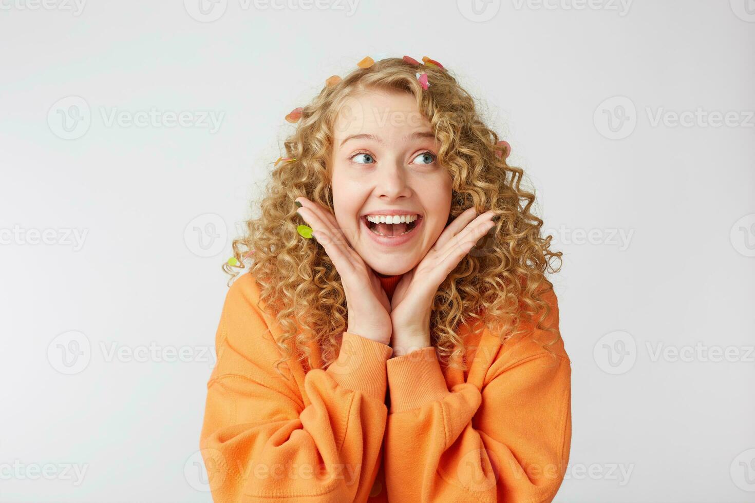 Close up of joyful smiling lovely tender nice blonde looks to the rightside feels excited, surprised, keeps palms near face, dressed in oversized orange sweater, isolated on a white background photo