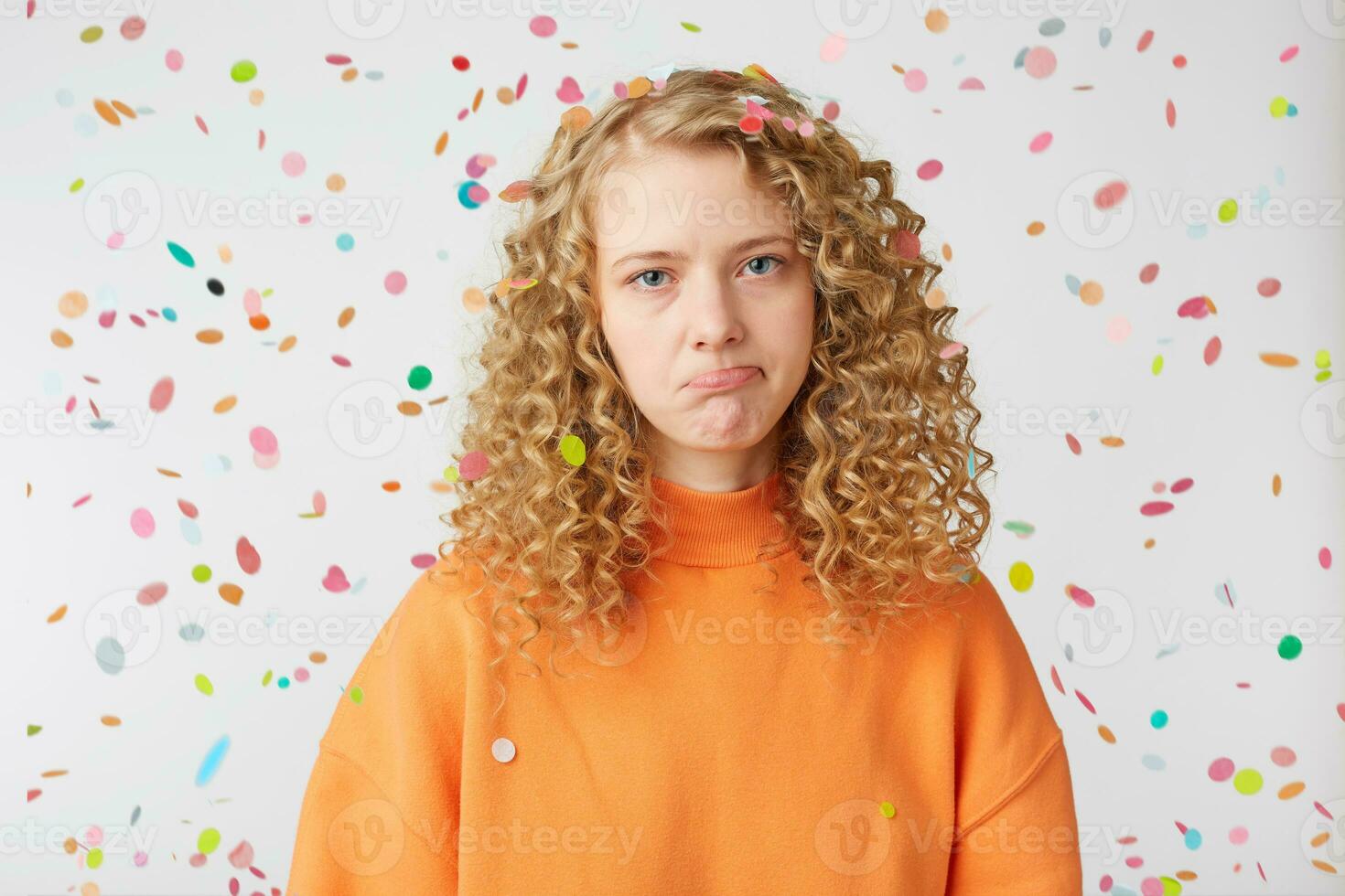 Close up of emotional girl looks sad, upset, frustrated, displeased, expresses offense,crying, lower lip pouted, showing defenseless, wears orange sweatshirt, stands under falling confetti. photo