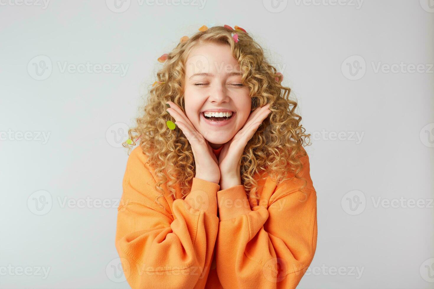 retrato de un alegre hermosa niña vistiendo naranja suéter mantiene palmas cerca cara celebrando con ojos cerrado en Placer aislado terminado blanco antecedentes foto